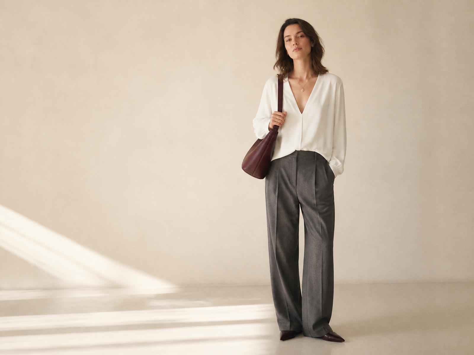 Warm white V-neck blouse worn with charcoal tailored trousers, a gold teardrop necklace and a burgundy shoulder bag